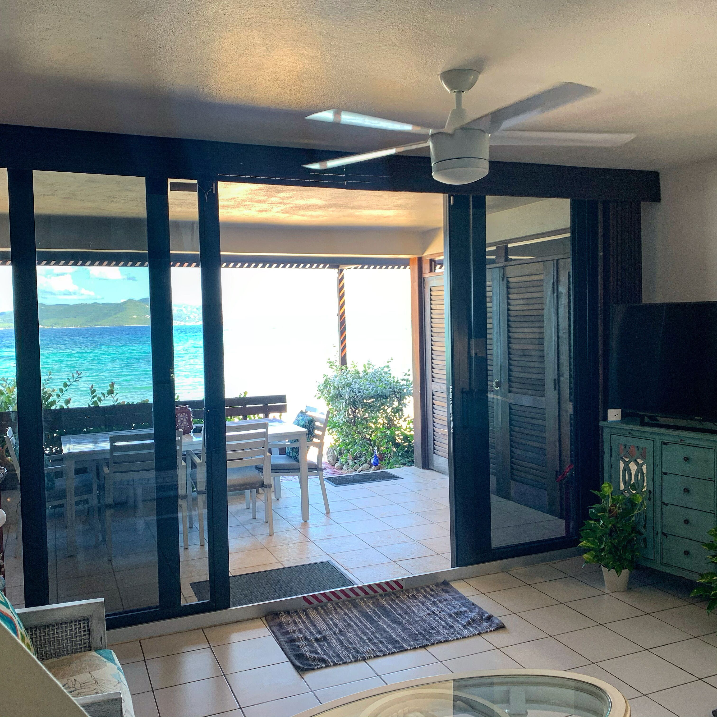 Een kamer met een schuifdeur die uitkomt op een balkon met uitzicht op de oceaan. Er is een plafondventilator, een televisie op een kast, en een eethoek met stoelen en een tafel.