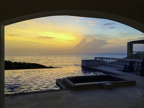 Vista a través de un ventanal arqueado sobre una piscina y el mar al atardecer.