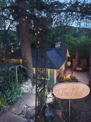 Un chalet rustique avec un sentier de pierres menant à une porte en bois, entouré d’arbres et illuminé de guirlandes lumineuses.