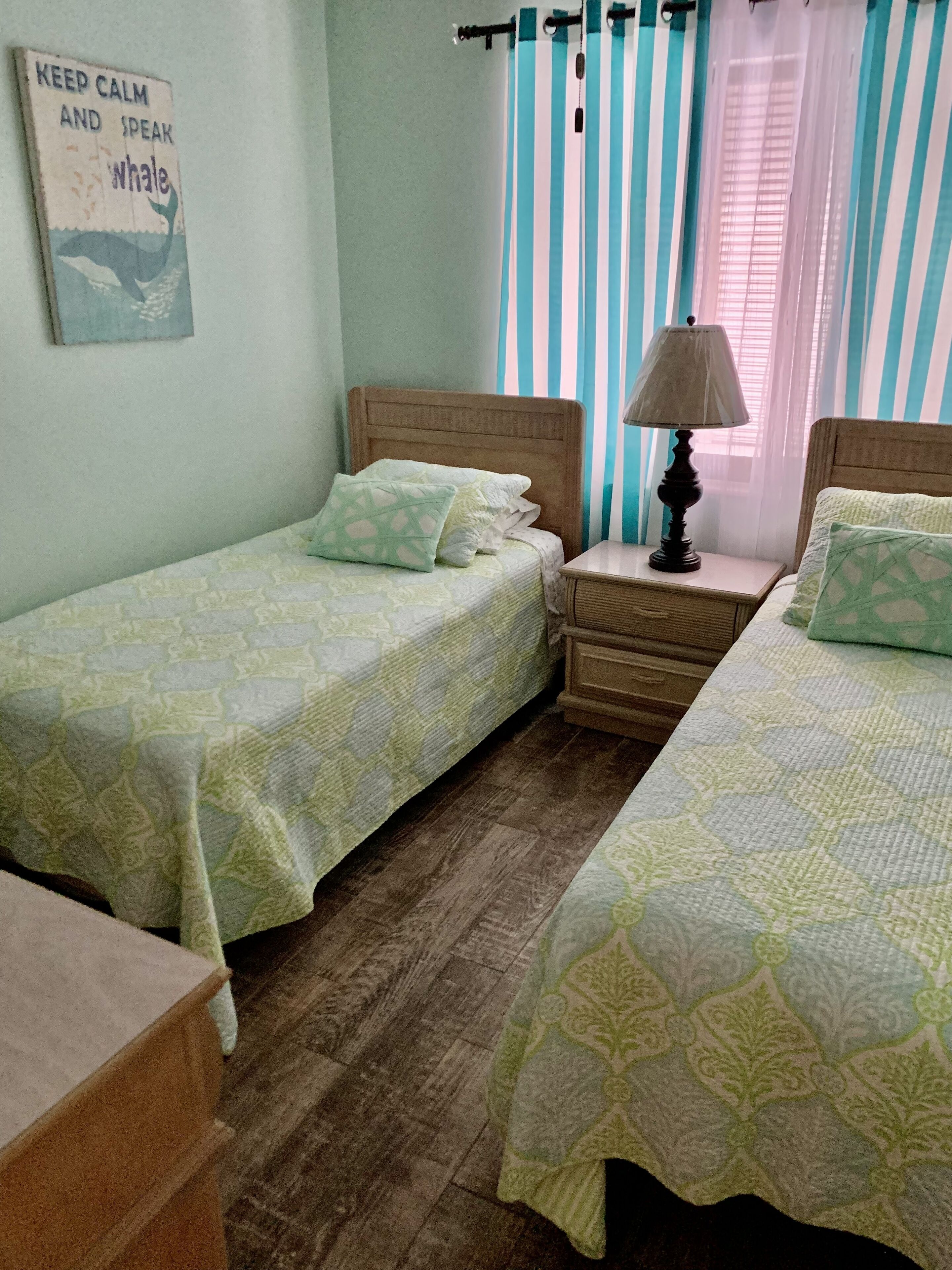 Une chambre avec deux lits simples, une table de chevet avec une lampe, des rideaux à rayures et un panneau décoratif mural.