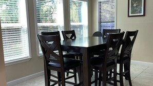 A dining area with a round table and six chairs, large windows with blinds, and a tiled floor.