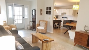 Un salon avec un canapé, une table basse en bois et une lampe. On aperçoit, au fond, une chaise en osier et une cuisine.