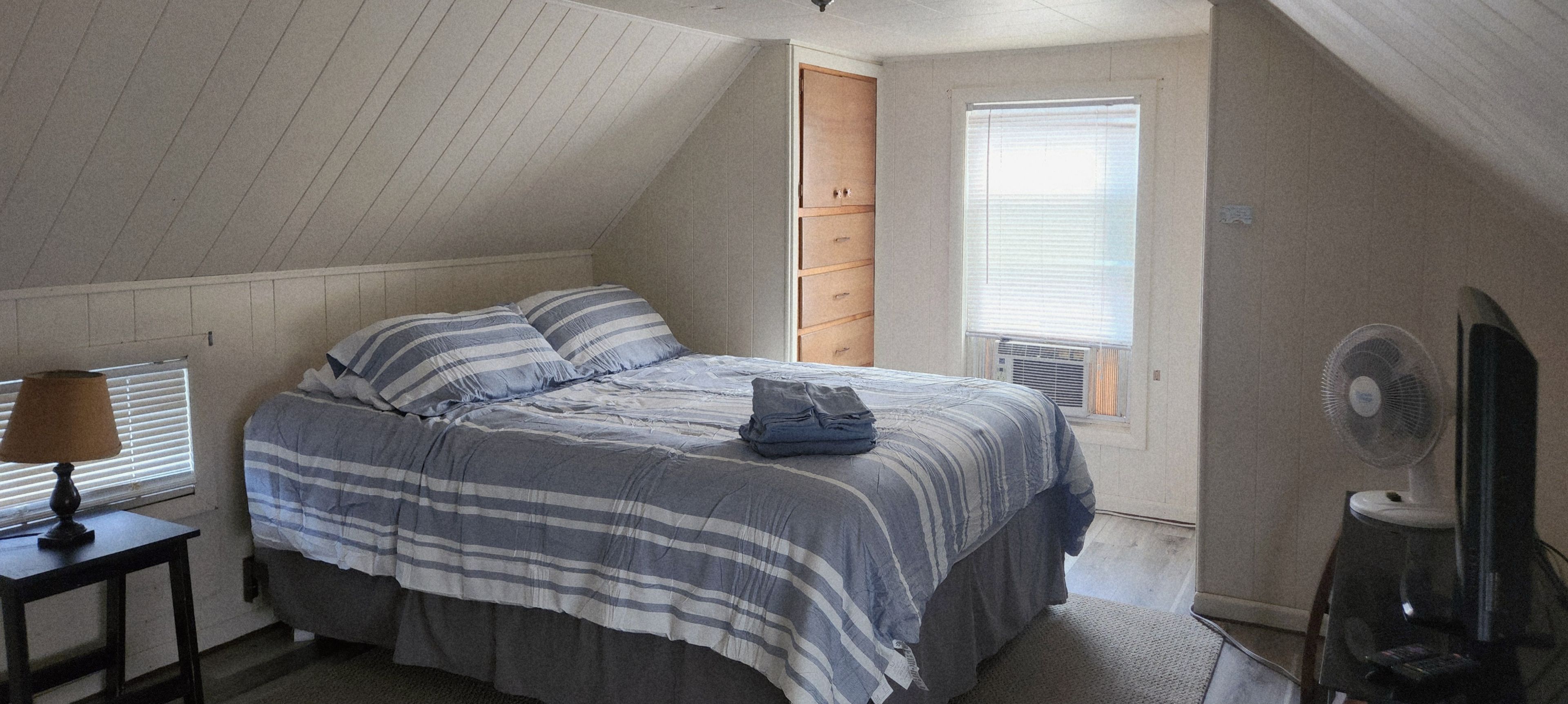 A bedroom with a sloped ceiling, a bed with striped bedding, a nightstand with a lamp, a dresser, and a window with blinds.