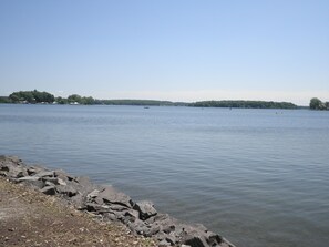 A calm lake with rocky shores and a distant shoreline with trees and buildings. - Amazing Lake View Cottage with Game room for Kids of all ages (Sodus Point)