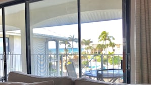 A balcony with a view of a pool and palm trees, a covered outdoor seating area, and a glass door leading to the room.
