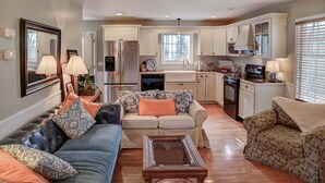 A spacious living room with a sofa, a coffee table, and a patterned armchair. A kitchen with white cabinets and a stainless steel refrigerator is visible in the background.