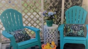 Zwei türkisblaue Stühle mit gemusterten Kissen, ein kleiner runder Tisch mit einem Blumentopf und ein Korb voller Gegenstände auf einer karierten Matte.