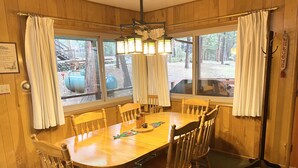A wooden dining room with a large table and chairs, large windows with curtains, and a chandelier.