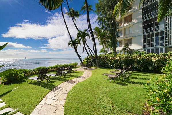 A well-maintained outdoor area with a stone pathway leading to lounge chairs and a table, overlooking a body of water with a sailboat in the distance.
