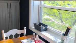 A dining area with a wooden table, white chairs, a window with blinds, and a view of greenery outside.