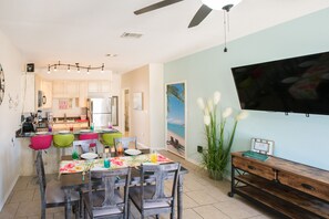 A modern kitchen with light-colored cabinets, a dining area with colorful chairs, and a large flat-screen TV mounted on the wall.