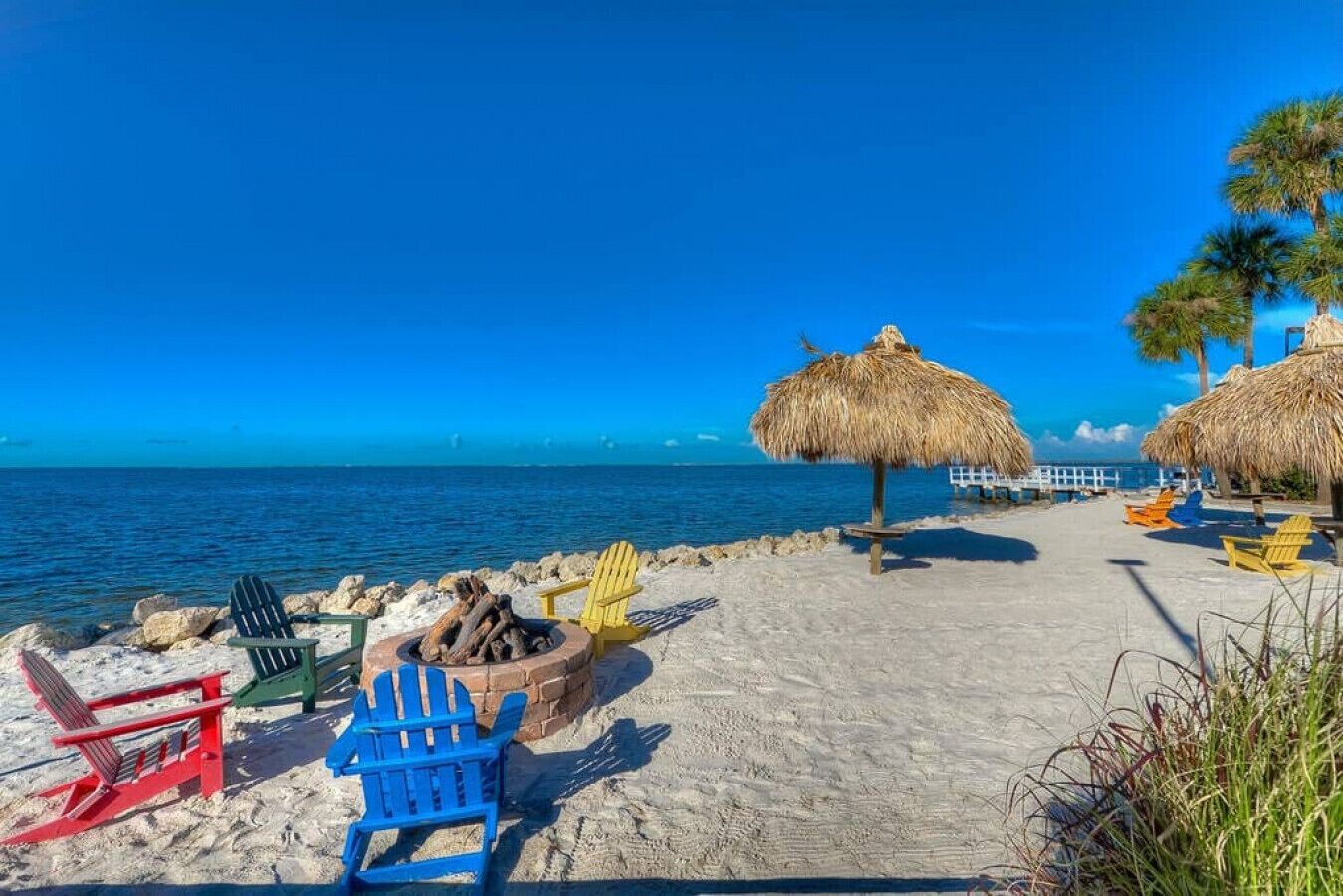 Une plage avec des parasols en chaume, des fauteuils Adirondack aux couleurs vives et un foyer.