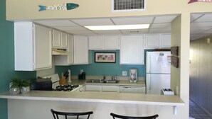Una cocina compacta con armarios blancos, estufa a gas, cafetera y dos taburetes de bar.