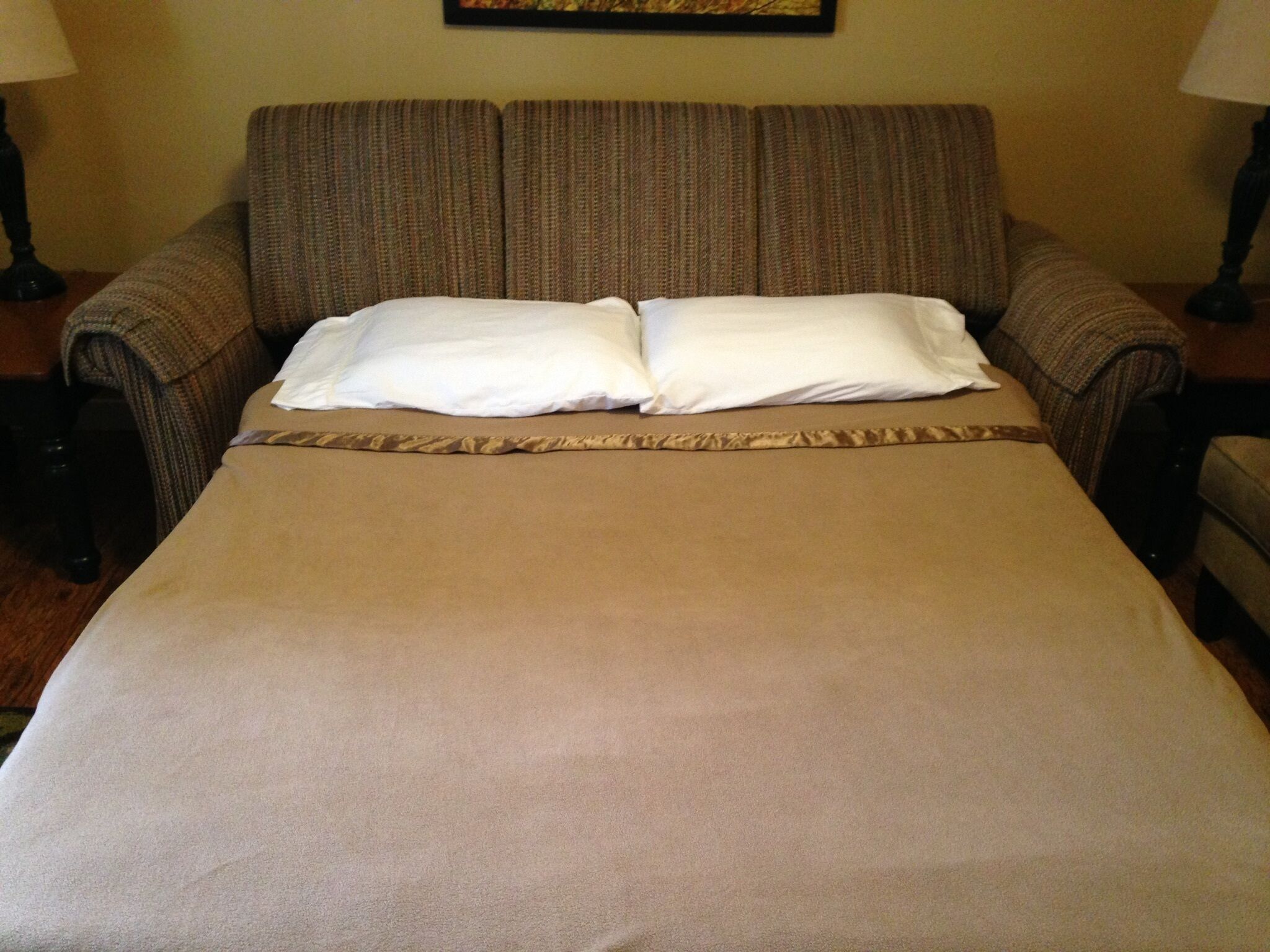 A bed with a beige bedspread and two pillows, flanked by a striped sofa and two nightstands with lamps.