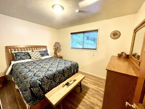 Un dormitorio con cama, un banco de madera, una mesita de noche, una lámpara, una ventana con persianas, un reloj y un espejo.