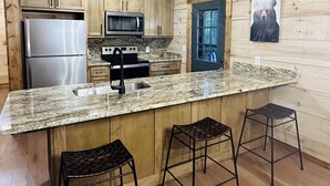 A kitchen with wooden cabinets, a stainless steel refrigerator, a microwave, and a granite countertop. There are two bar stools and a window with a bear picture.