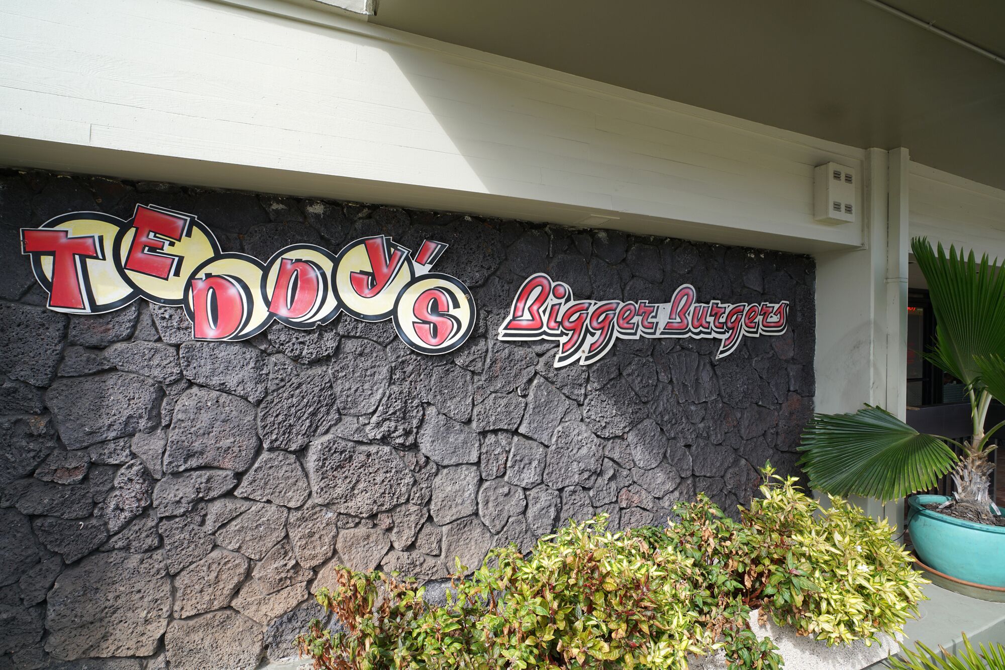 Une enseigne pour « Tendo’s Biger Burgers », avec un mur de pierres en arrière-plan et quelques plantes vertes devant.