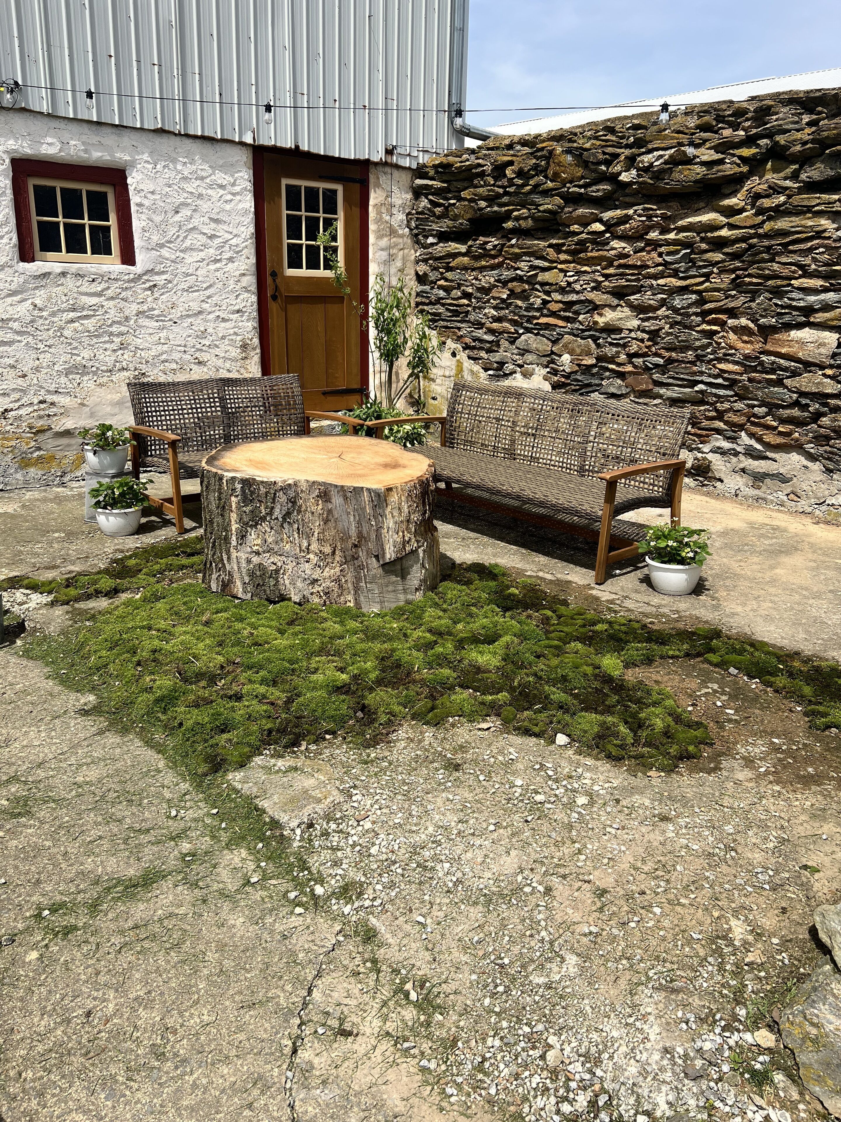 A rustic outdoor seating area with a round wooden table and wicker chairs, surrounded by potted plants and a stone wall.