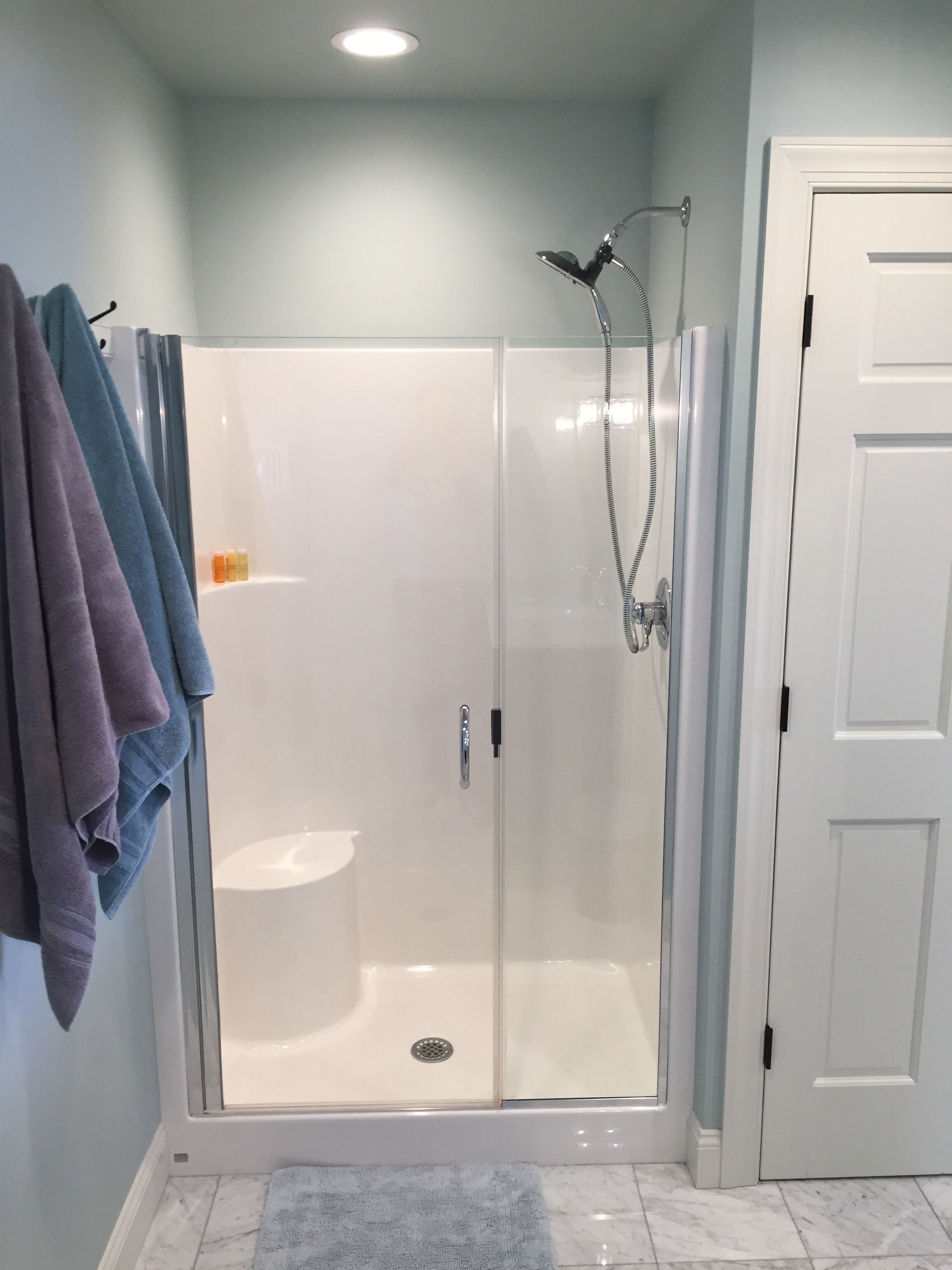A glass-enclosed shower stall with a handheld showerhead, a white door, and a towel rack with towels.