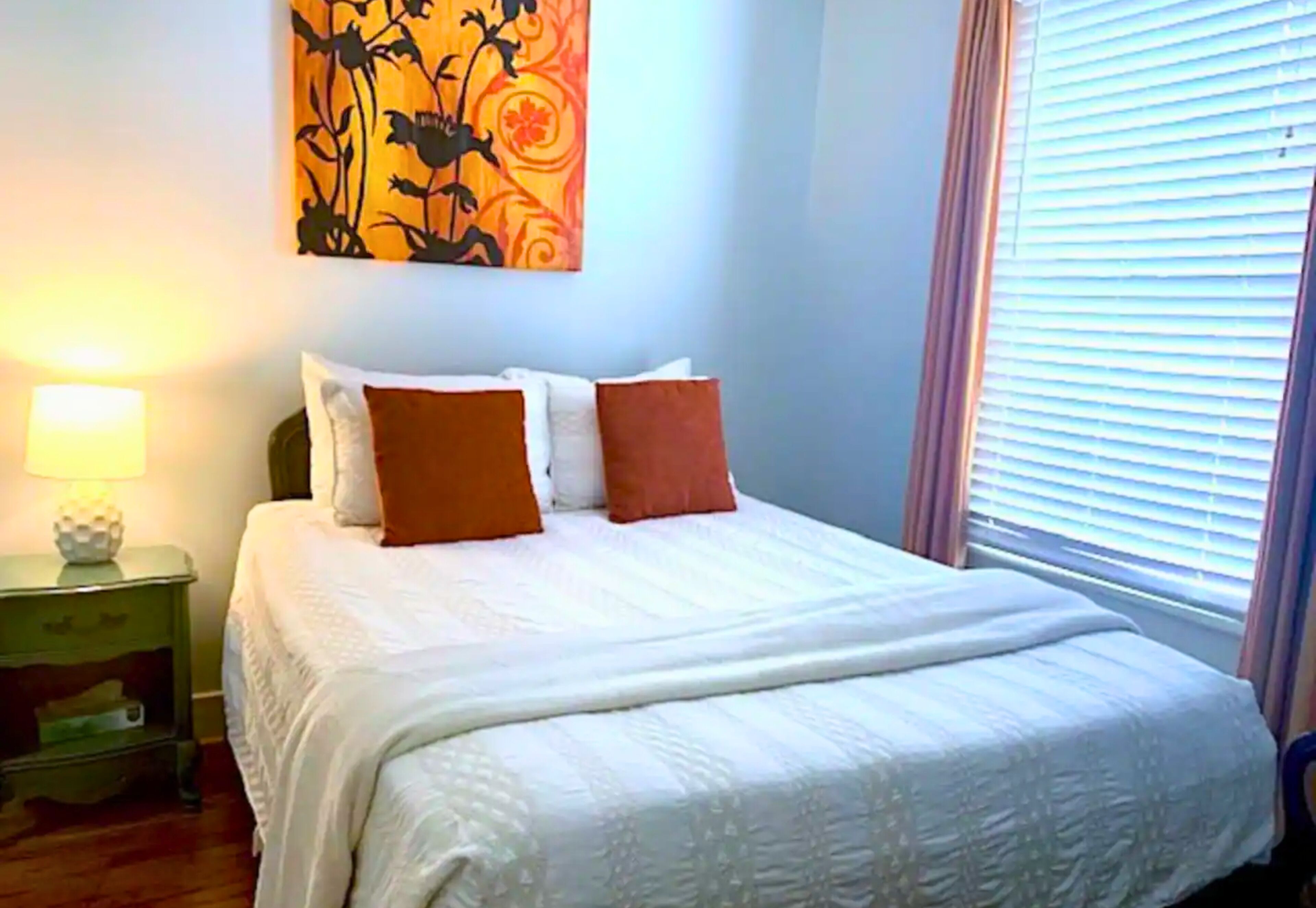 Een net opgemaakt bed met wit en oranje beddengoed, een kunstwerk erboven en een raam met jaloezieën aan de rechterkant.