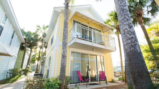 A two-story house with a balcony, surrounded by palm trees and a small garden.