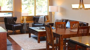 A living room with a wooden dining table, chairs, a sofa, and a coffee table. There are two windows with blinds, a lamp, and a chandelier. - Mountain + Creek View. 5 Min Walk To Main Street. An Amazing Home Base! (Canmore)