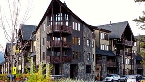 A multi-story lodge with a stone facade, wooden balconies, and a ski lift visible in the background. - Mountain + Creek View. 5 Min Walk To Main Street. An Amazing Home Base! (Canmore)