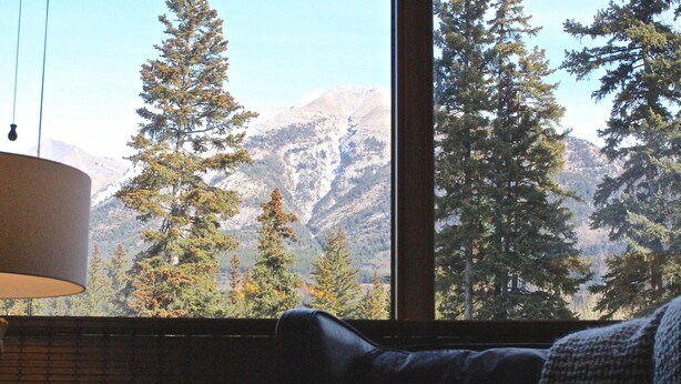 A room with a large window offering a view of a mountainous landscape with evergreen trees. - Mountain + Creek View. 5 Min Walk To Main Street. An Amazing Home Base! (Canmore)