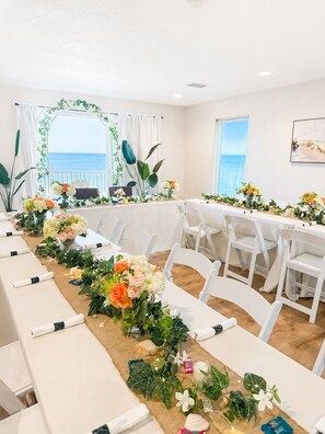 Una larga mesa con sillas blancas, decorada con flores y follaje, en un espacio con vistas al mar.
