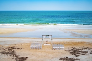 Hochzeitsdekoration am Strand mit weißen Stühlen und einem hölzernen Torbogen.