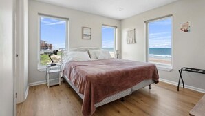 A bedroom with a bed, a nightstand, a desk, and a bench. There are large windows with a view of the beach and ocean.