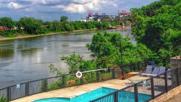 Una piscina con vista a un río y estructuras industriales al fondo.