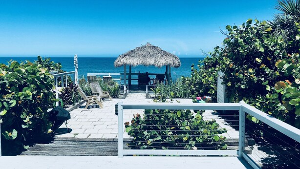 A beachside walkway leading to a thatched-roof structure with outdoor seating and a view of the ocean.