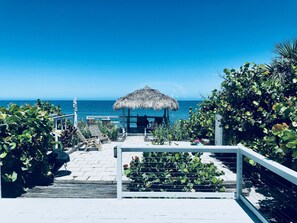 A beachside walkway leading to a thatched-roof structure with outdoor seating and a view of the ocean. - Secluded Tiki Beach House - Beach fun, surf, meditate, fish, play or just relax (New Smyrna Beach)