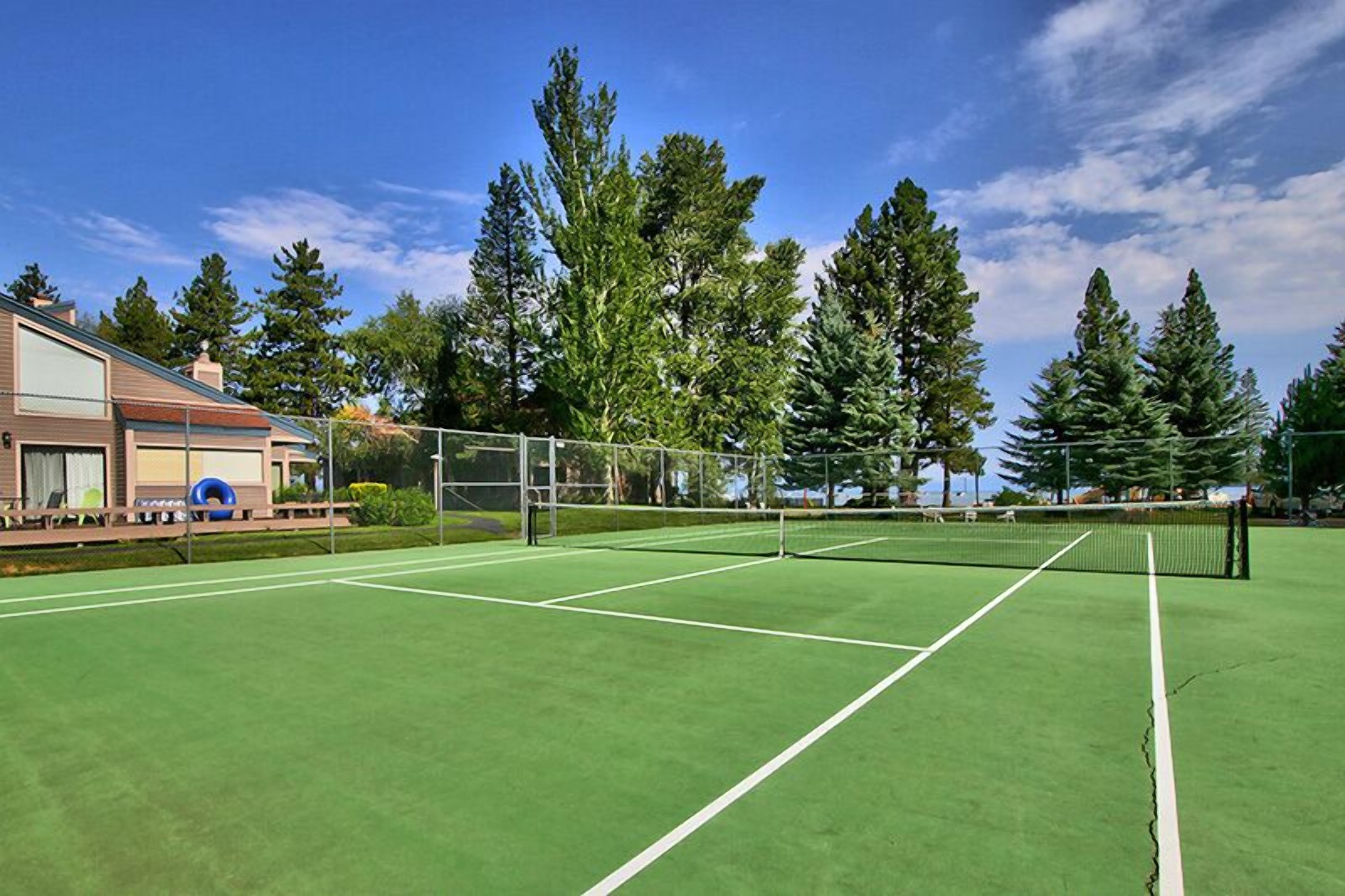 Ein Tennisplatz mit Netz, umgeben von Bäumen und einem Gebäude mit überdachter Sitzgelegenheit.