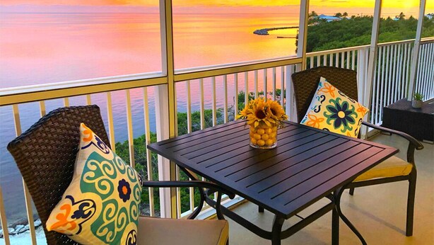 Un balcone con tavolo e sedie, un vaso con girasoli e vista sull'oceano al tramonto.