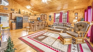 A cozy log cabin interior with a wooden ceiling, a kitchen area, a dining table with chairs, a sofa, and a fireplace. - H1 - Mountain Cabin-Hot Tub-1.5 miles to Dollywood-Resort Pool-No Mountain Roads (Sevierville)