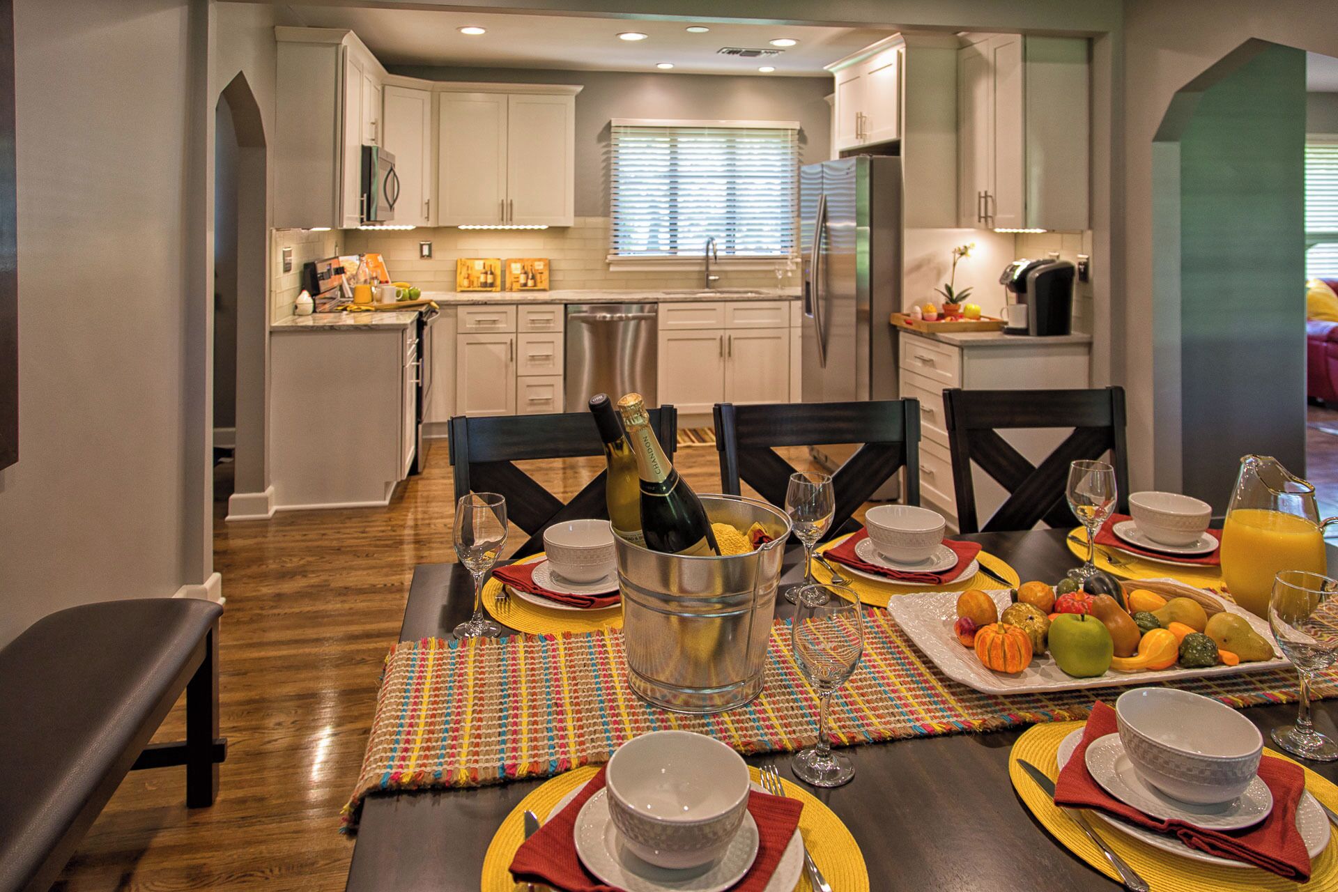 Comedor con una mesa lista para comer, cocina al fondo y una ventana con persianas.