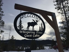 Señal circular con silueta de alce y el texto 'Welcome Whispering Pines Guest Cabin'.
