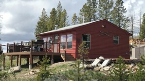 A red cabin with a deck, surrounded by trees and a clear sky.