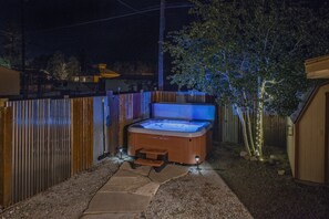 Un bain à remous dans un jardin, avec une clôture en bois, une terrasse en bois et un arbre décoré de guirlandes lumineuses.