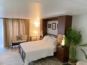 A hotel room with a bed, a chair, a nightstand, a lamp, and a potted plant.