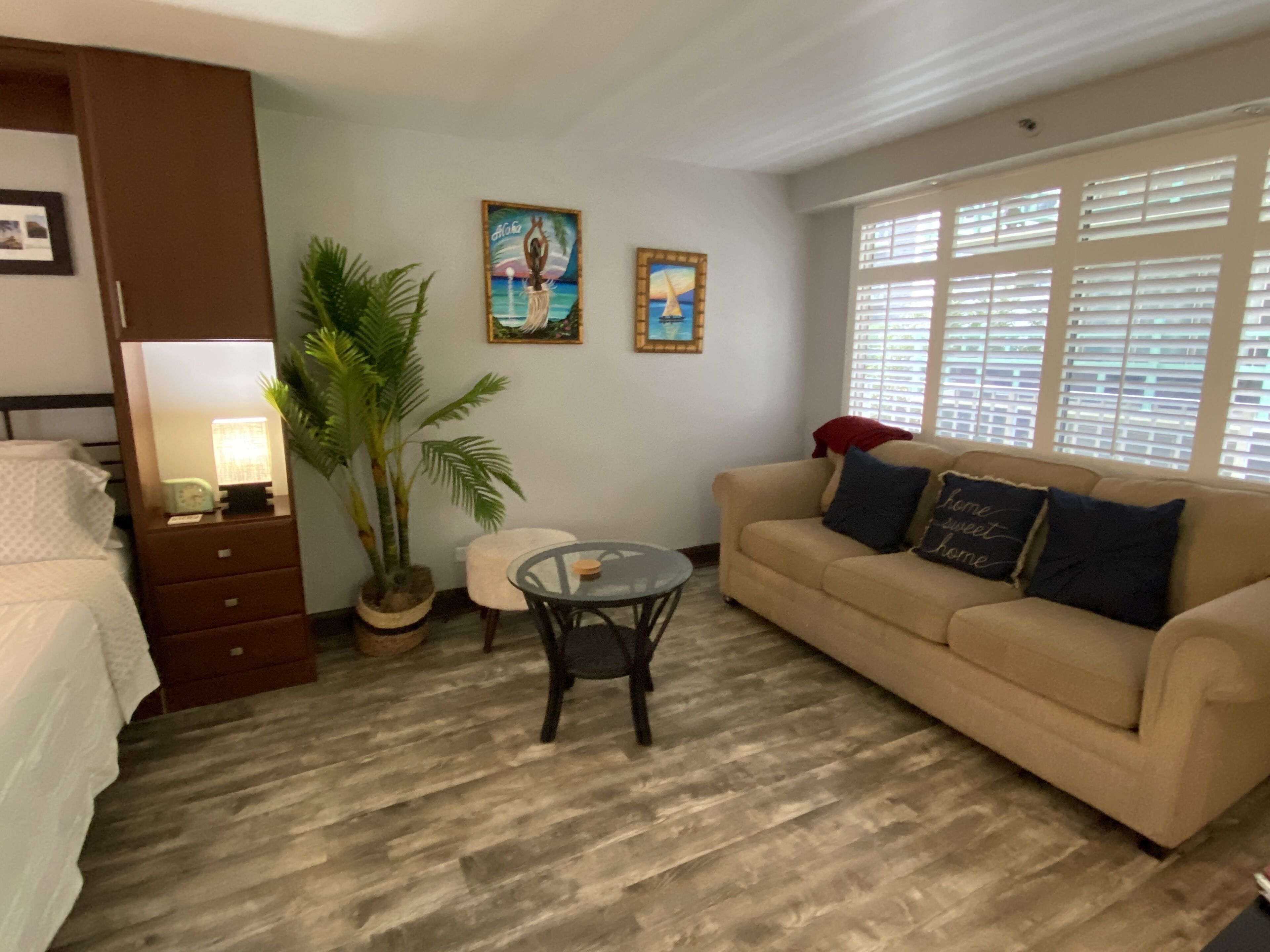 A room with a bed, a sofa, a small table, a potted plant, and two wall-mounted paintings.