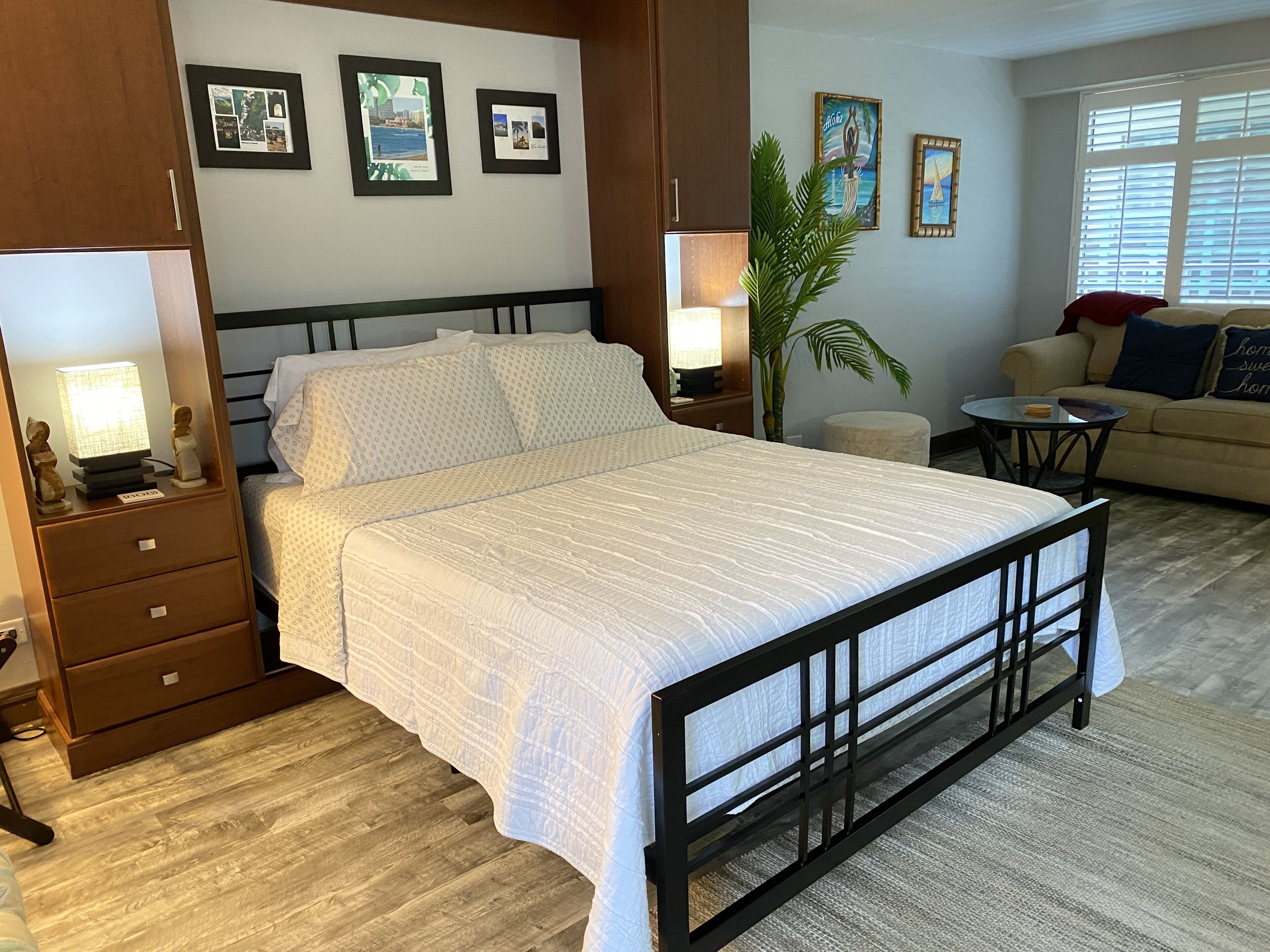 A bedroom with a bed, bedside tables, a nightstand, a lamp, a plant, a sofa, and framed pictures on the wall.