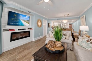 A spacious living room with a flat-screen TV, a fireplace, a coffee table with decorative items, a sofa, and a dining area in the background.