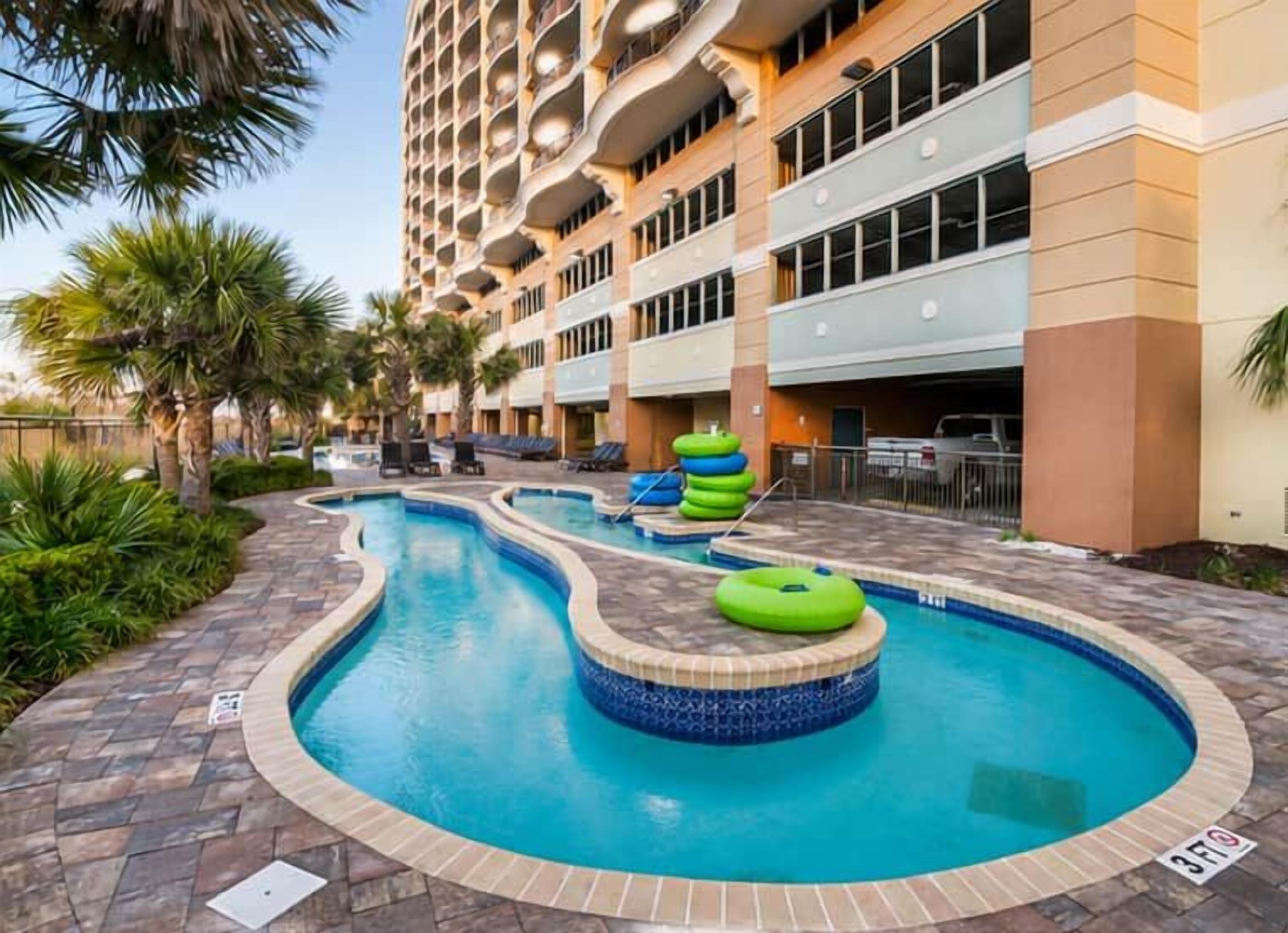 A hotel pool area with multiple swimming pools, a hot tub, and a parking garage.