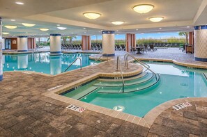 A multi-level indoor swimming pool with clear water, surrounded by lounge chairs and sunbeds, and a view of the outdoors.
