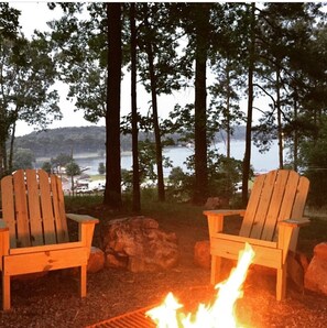Two Adirondack chairs facing a campfire with a lake and forest in the background.
