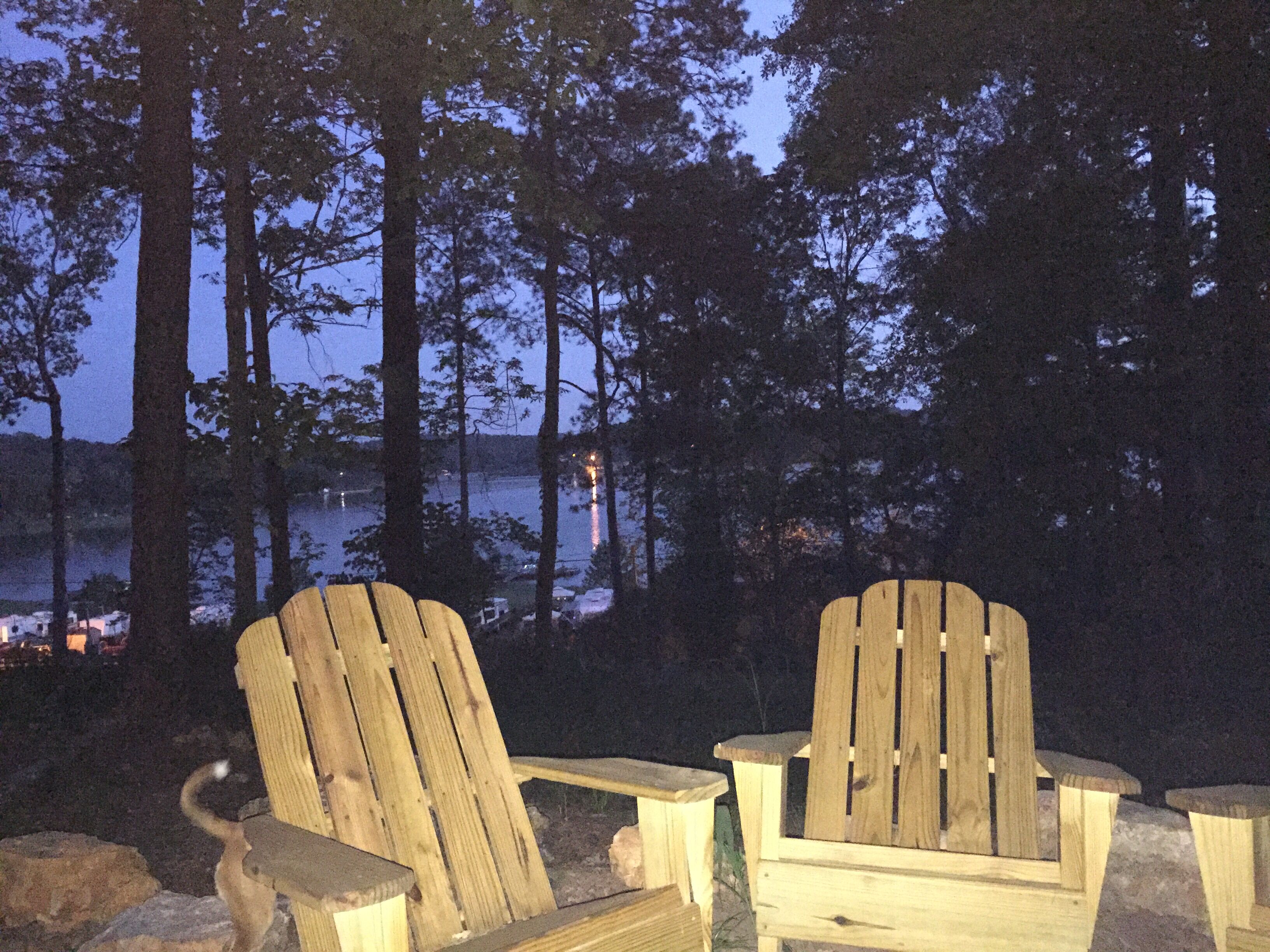 Deux fauteuils Adirondack en bois, placés face à une étendue d’eau, avec des arbres en arrière-plan.