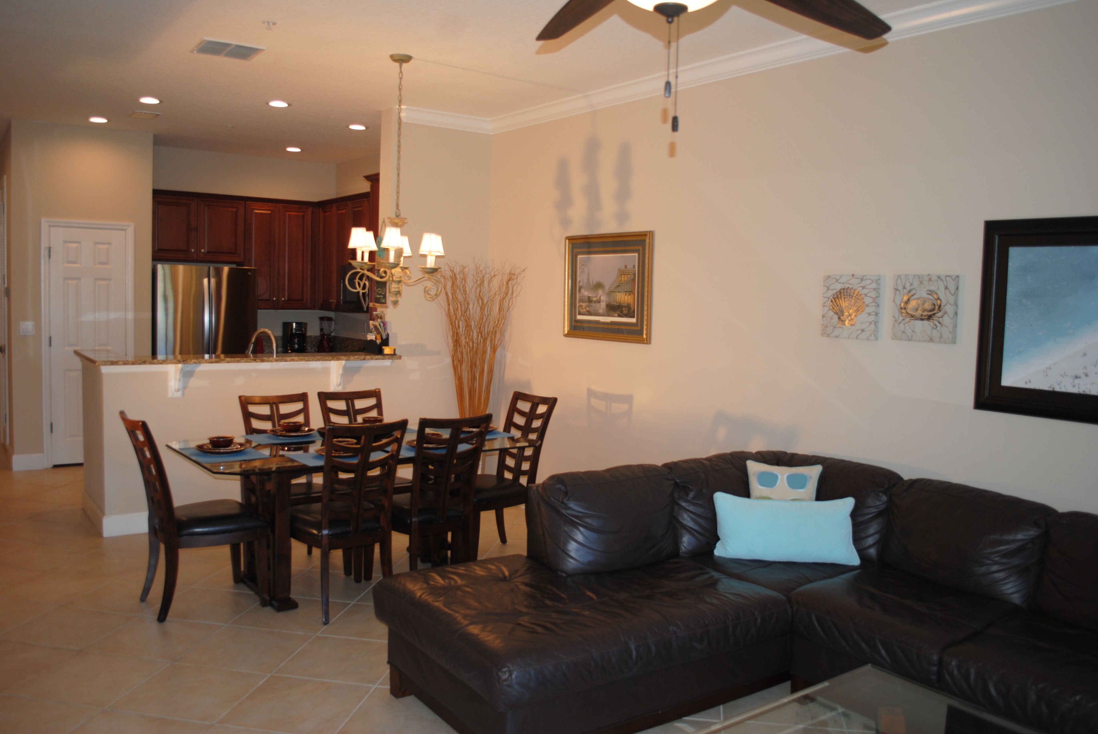 A living room with a black leather sofa, a dining table with chairs, a kitchen area with cabinets, and a ceiling fan.